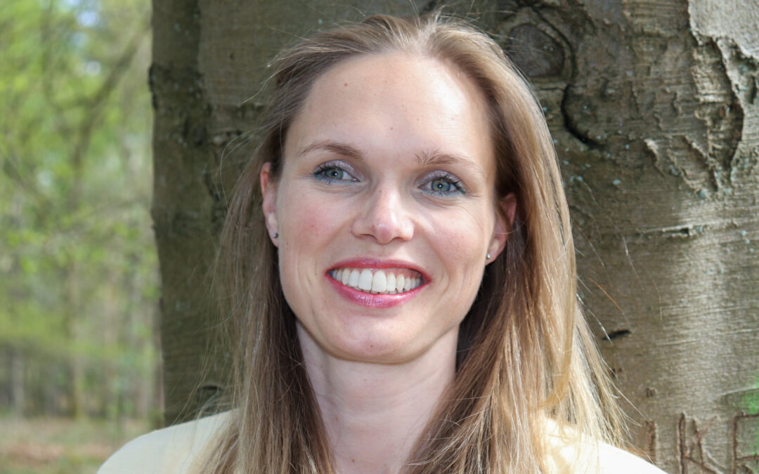 Tjarda staat buiten en draagt een licht gele trui. Ze lacht en kijkt in de camera.