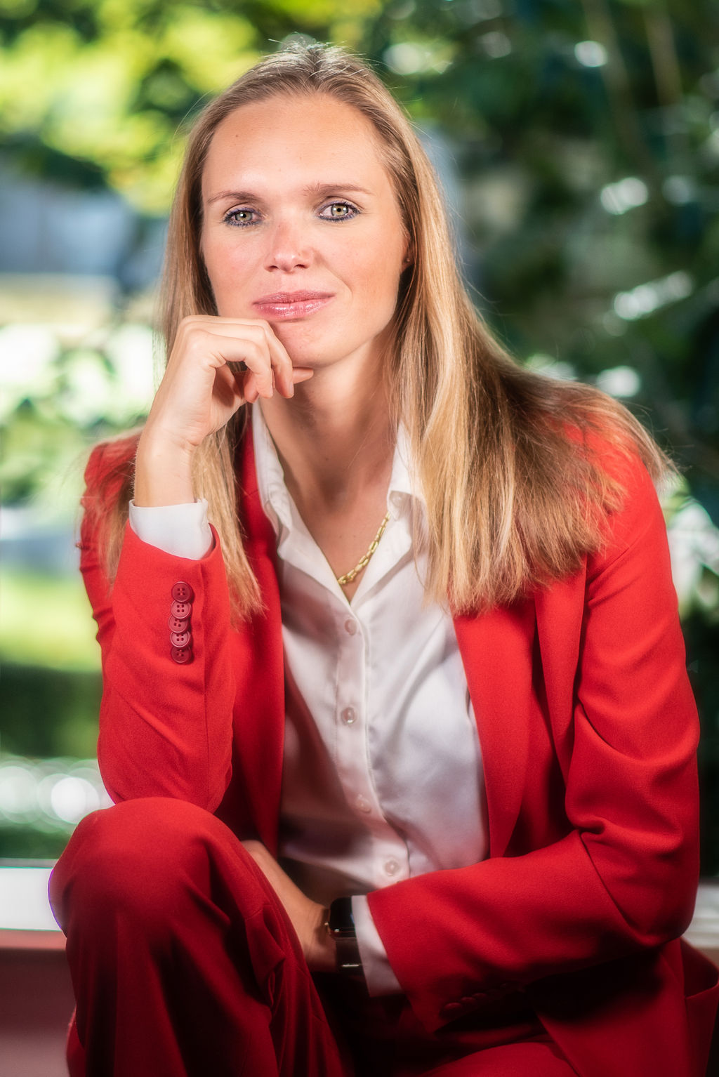 Tjarda zit in een raamkozijn en draagt een blauwe blazer. Haar ene arm rust op haar knie en haar andere arm langs haar zijde.